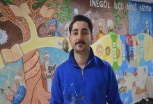 Renkli bir duvar resminin önünde duran bir kişi kameraya doğru bakıyor. Üzerinde boya lekeleri olan mavi bir üst var. Arka planda eğitim temalı figürler, kitaplar ve tarihi karakterlerin yer aldığı detaylı bir mural bulunuyor. Sahne, sanatsal bir çalışmanın ortasında verilen bir röportaj ya da tanıtım anını yansıtıyor.