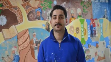 Renkli bir duvar resminin önünde duran bir kişi kameraya doğru bakıyor. Üzerinde boya lekeleri olan mavi bir üst var. Arka planda eğitim temalı figürler, kitaplar ve tarihi karakterlerin yer aldığı detaylı bir mural bulunuyor. Sahne, sanatsal bir çalışmanın ortasında verilen bir röportaj ya da tanıtım anını yansıtıyor.