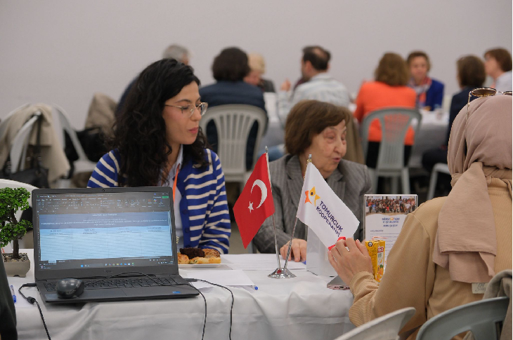 Görselde bir çalıştay ortamında masada karşılıklı oturan katılımcılar görülüyor. Masada bilgisayar, belgeler ve küçük masa bayrakları (Türkiye bayrağı ve bir dernek/kurum bayrağı) bulunuyor. Katılımcılar tartışma veya değerlendirme yaparken, arka planda diğer grupların da benzer şekilde çalıştığı bir toplantı salonu görülüyor. Ortam, yoğun ve iş birlikçi bir çalışma atmosferi yansıtıyor.