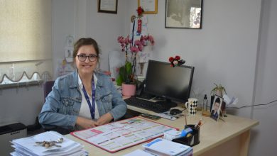 Görselde, bir ofis ortamında masasında oturan ve gülümseyen bir kadın görülüyor. Masanın üzerinde takvim, belgeler, kalemlik, bilgisayar ve çeşitli kişisel eşyalar yer alıyor. Arka planda çerçeveli belgeler ve çiçekler bulunuyor. Ortam sakin ve düzenli bir çalışma alanı izlenimi veriyor.