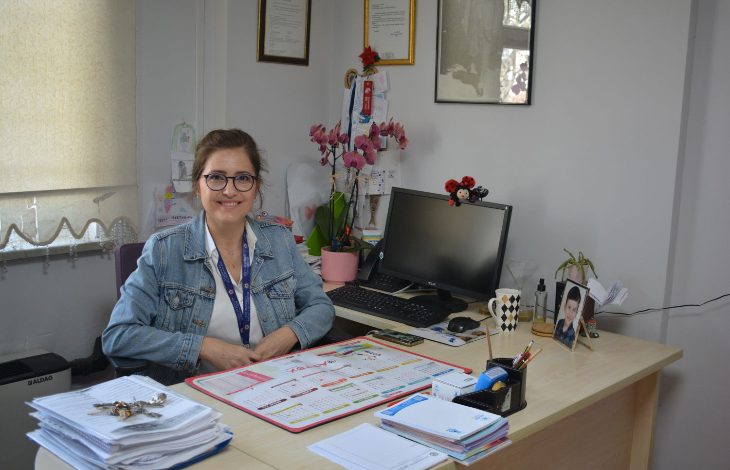 Görselde, bir ofis ortamında masasında oturan ve gülümseyen bir kadın görülüyor. Masanın üzerinde takvim, belgeler, kalemlik, bilgisayar ve çeşitli kişisel eşyalar yer alıyor. Arka planda çerçeveli belgeler ve çiçekler bulunuyor. Ortam sakin ve düzenli bir çalışma alanı izlenimi veriyor.