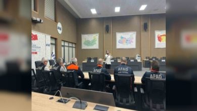 Bir toplantı salonunda AFAD personelleri oturmuş, ön tarafta ayakta duran bir kişi sunum yapıyor. Duvarlarda haritalar asılı; ortam resmi bir eğitim ya da bilgilendirme toplantısını andırıyor.