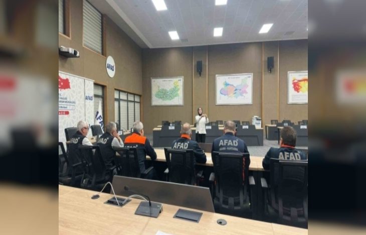 Bir toplantı salonunda AFAD personelleri oturmuş, ön tarafta ayakta duran bir kişi sunum yapıyor. Duvarlarda haritalar asılı; ortam resmi bir eğitim ya da bilgilendirme toplantısını andırıyor.