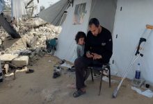 Görselde, yıkıntıların yanında kurulu bir çadırın önünde oturan bir adam, elindeki telefona bakarken yanında küçük bir çocuk sessizce ona eşlik ediyor; çevredeki dağınıklık ve bastonlar zor koşulları düşündürüyor.