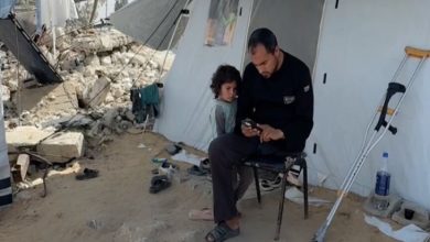 Görselde, yıkıntıların yanında kurulu bir çadırın önünde oturan bir adam, elindeki telefona bakarken yanında küçük bir çocuk sessizce ona eşlik ediyor; çevredeki dağınıklık ve bastonlar zor koşulları düşündürüyor.