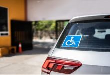 Bir aracın arka camında engelli park kartı işareti bulunuyor. Araç bir bina girişinin önünde park edilmiş; çevrede kapı ve güvenlik alanı görülüyor.