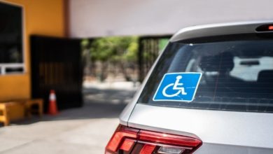 Bir aracın arka camında engelli park kartı işareti bulunuyor. Araç bir bina girişinin önünde park edilmiş; çevrede kapı ve güvenlik alanı görülüyor.