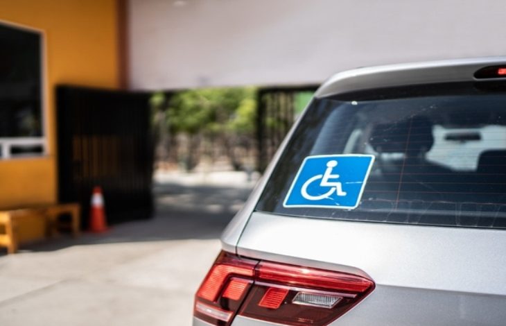 Bir aracın arka camında engelli park kartı işareti bulunuyor. Araç bir bina girişinin önünde park edilmiş; çevrede kapı ve güvenlik alanı görülüyor.