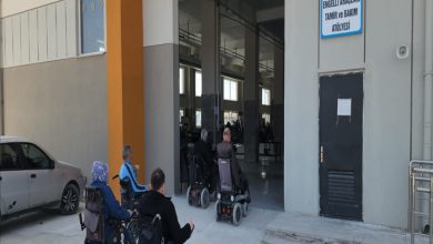 Tekerlekli sandalye kullanan bir grup bireyin, üzerinde "Engelli Araçları Tamir ve Bakım Atölyesi" tabelası bulunan bir binaya giriş yaptığı anı gösteren görsel. Fotoğraf, engelli bireylerin hareketliliğini destekleyen bir hizmet merkezini ve bu merkeze olan erişimi yansıtıyor.