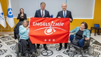Görselde, İzmir Büyükşehir Belediye Başkanı Dr. Cemil Tugay, İZTO Yönetim Kurulu Başkanı Mahmut Özgener ile iki tekerlekli sandalye kullanan kadının birlikte “Engelsiz İzmir” yazılı kırmızı bir pankart tutarak engelli bireyler için farkındalık mesajı verdiği bir etkinlik anı görülüyor.