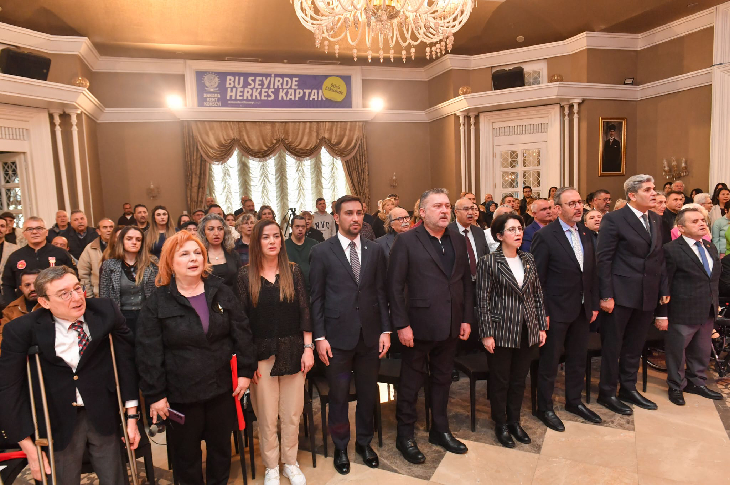 Görselde, bir salon toplantısında resmi bir etkinlik için ayakta duran kalabalık bir grup görülüyor. Ön sıradaki katılımcılar protokolü andıran bir düzende dizilmiş durumda. Arkada ise etkinliği takip eden geniş bir izleyici kitlesi yer alıyor. Salon avizesi, perde detayları ve duvardaki portre ile birlikte klasik bir toplantı salonu atmosferi yansıtıyor. Genel görünüm, kurumsal bir toplantı veya genel kurul oturumuna işaret ediyor.