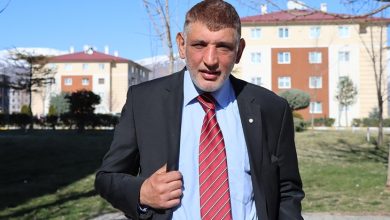 Açık havada çekilmiş bir portre görülüyor. Takım elbise ve kırmızı çizgili kravat takan orta yaşlı bir erkek, yürüyüş yolunda kameraya doğru ilerlerken ceketini tutuyor. Arka planda apartmanlar, ağaçlar ve güneşli bir günün net ışığı var. Yüz ifadesi ciddi ama kararlı bir duruş yansıtıyor.