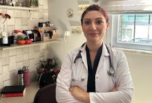 Görselde, beyaz önlük giymiş bir doktor muayene odasında ayakta duruyor. Boynunda stetoskopu var ve kollarını çaprazlamış şekilde gülümseyerek poz veriyor. Arka planda masa, raflar ve bir pencere görülüyor.