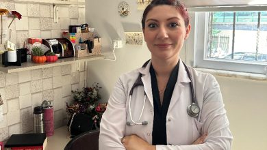 Görselde, beyaz önlük giymiş bir doktor muayene odasında ayakta duruyor. Boynunda stetoskopu var ve kollarını çaprazlamış şekilde gülümseyerek poz veriyor. Arka planda masa, raflar ve bir pencere görülüyor.