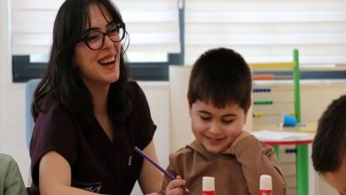 Bir sınıf ortamında, gözlüklü bir kadın öğretmen küçük bir çocukla birlikte masa başında etkinlik yapıyor. Kadın gülerek çocuğa eşlik ederken, çocuk elinde kalemle çalışıyor ve mutlu görünüyor. Masada yapıştırıcılar var; arka planda renkli eğitim materyalleri ve başka bir çocuk daha kısmen görünüyor. Atmosfer sıcak ve destekleyici bir öğrenme anını yansıtıyor.