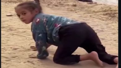 Kumlu bir zeminde, küçük bir çocuk emekler halde ilerliyor. Başını geriye çevirip kameraya bakıyor. Üzerinde uzun kollu bir üst ve koyu renk pantolon var; arka planda bulanık şekilde başka biri görülüyor.