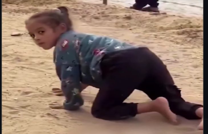 Kumlu bir zeminde, küçük bir çocuk emekler halde ilerliyor. Başını geriye çevirip kameraya bakıyor. Üzerinde uzun kollu bir üst ve koyu renk pantolon var; arka planda bulanık şekilde başka biri görülüyor.