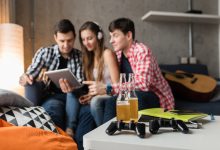 Ev ortamında üç genç birlikte oturmuş, bir tablet ekranına odaklanmış halde vakit geçiriyor. Ön planda masada içecek şişeleri, oyun kumandası ve defterler var; arka planda ise bir gitar dikkat çekiyor. Sahne, sosyal ama aynı zamanda dijital odaklı bir anı yansıtıyor.