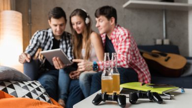 Ev ortamında üç genç birlikte oturmuş, bir tablet ekranına odaklanmış halde vakit geçiriyor. Ön planda masada içecek şişeleri, oyun kumandası ve defterler var; arka planda ise bir gitar dikkat çekiyor. Sahne, sosyal ama aynı zamanda dijital odaklı bir anı yansıtıyor.