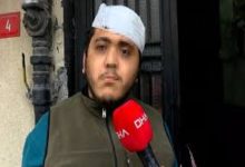 Sokakta bir binanın girişinde duran genç bir adam, elindeki kırmızı mikrofonla röportaj veriyor. Başında bandaj bulunuyor. Arka planda bina kapısı ve duvar detayları görülüyor. Sahne, yaşadığı bir olay sonrası yapılan bir açıklama anını yansıtıyor.