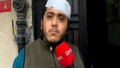 Sokakta bir binanın girişinde duran genç bir adam, elindeki kırmızı mikrofonla röportaj veriyor. Başında bandaj bulunuyor. Arka planda bina kapısı ve duvar detayları görülüyor. Sahne, yaşadığı bir olay sonrası yapılan bir açıklama anını yansıtıyor.