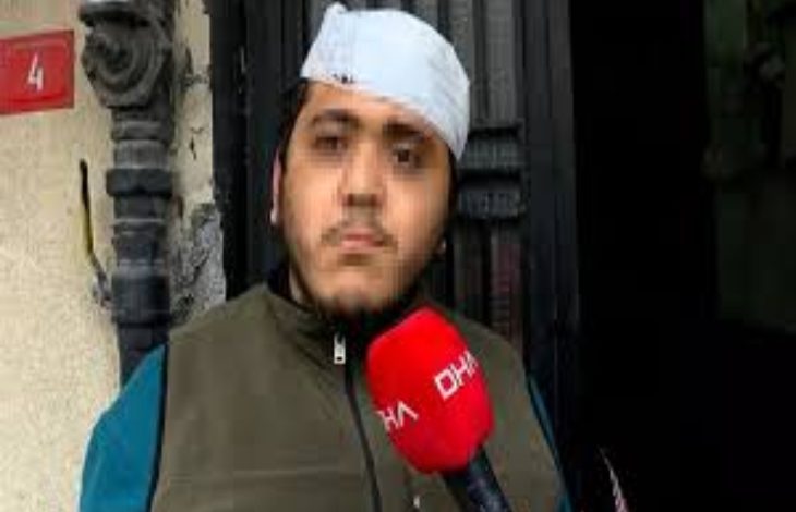 Sokakta bir binanın girişinde duran genç bir adam, elindeki kırmızı mikrofonla röportaj veriyor. Başında bandaj bulunuyor. Arka planda bina kapısı ve duvar detayları görülüyor. Sahne, yaşadığı bir olay sonrası yapılan bir açıklama anını yansıtıyor.