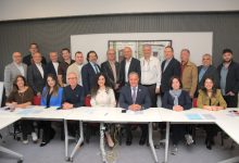 Geniş bir katılımcı grubunun bir toplantı salonunda, beyaz masaların etrafında ve arkasında toplu halde verdiği resmî bir heyet fotoğrafı. Katılımcılar profesyonel giyimli, ortam ise kurumsal bir toplantı atmosferini yansıtıyor.