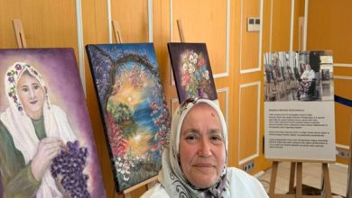 Bir sergi alanında başörtülü bir kadın kameraya bakıyor; arkasında şövalelere yerleştirilmiş renkli tablolar (portre ve doğa temalı) sergileniyor. Sağ tarafta eserlerle ilgili açıklama panosu bulunuyor; ortam küçük bir sanat sergisini andırıyor.