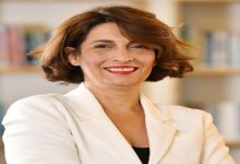 Kısa saçlı, bakır tonlu bir kadın; açık renk ceketle, kolları çapraz şekilde poz veriyor. Gülümseyen, kendinden emin bir ifade ve arka planda bulanık bir kitaplık görülüyor.