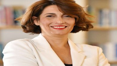 Kısa saçlı, bakır tonlu bir kadın; açık renk ceketle, kolları çapraz şekilde poz veriyor. Gülümseyen, kendinden emin bir ifade ve arka planda bulanık bir kitaplık görülüyor.