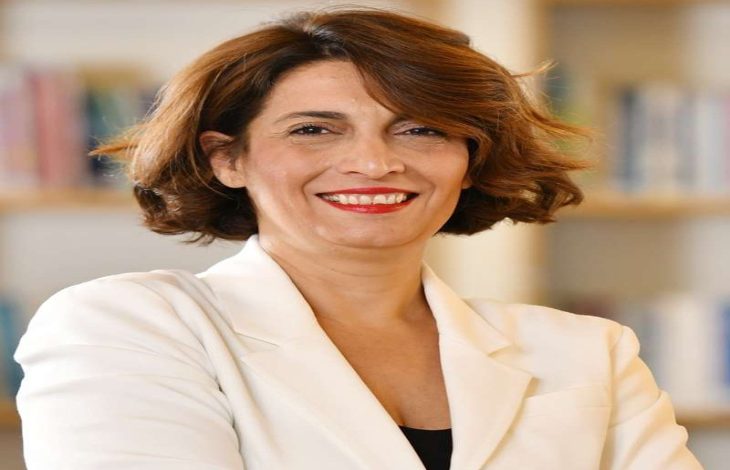 Kısa saçlı, bakır tonlu bir kadın; açık renk ceketle, kolları çapraz şekilde poz veriyor. Gülümseyen, kendinden emin bir ifade ve arka planda bulanık bir kitaplık görülüyor.