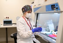 Laboratuvar ortamında maske, eldiven ve önlük giymiş kadın sağlık çalışanı, steril bir kabin içinde numune tüpüyle dikkatlice çalışıyor.