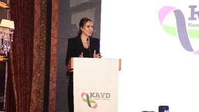 Görselde bir kadın, kürsüde konuşma yaparken görülüyor. Siyah bir takım elbise giymiş, saçları toplu ve mikrofon aracılığıyla dinleyicilere hitap ediyor. Kürsünün üzerinde bir dernek logosu yer alıyor. Arka planda büyük bir ekran ve sahne düzeni bulunuyor. Ön tarafta izleyicilerden bazıları konuşmayı telefonlarıyla kaydediyor.