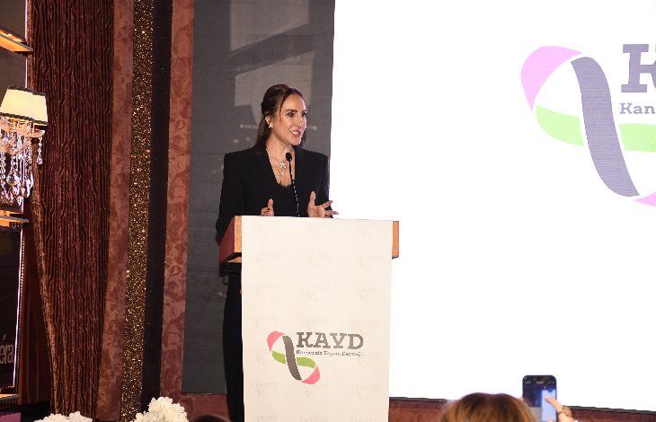 Görselde bir kadın, kürsüde konuşma yaparken görülüyor. Siyah bir takım elbise giymiş, saçları toplu ve mikrofon aracılığıyla dinleyicilere hitap ediyor. Kürsünün üzerinde bir dernek logosu yer alıyor. Arka planda büyük bir ekran ve sahne düzeni bulunuyor. Ön tarafta izleyicilerden bazıları konuşmayı telefonlarıyla kaydediyor.