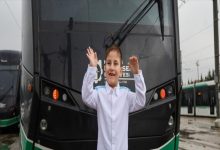 Yağmurlu bir günde, tramvayın önünde duran küçük bir çocuk iki elini havaya kaldırmış, gülümseyerek poz veriyor. Üzerinde beyaz bir gömlek var. Arkada raylı sistem araçları ve şehir altyapısı görünüyor; sahne hem neşeli hem umut dolu bir anı yakalamış gibi.