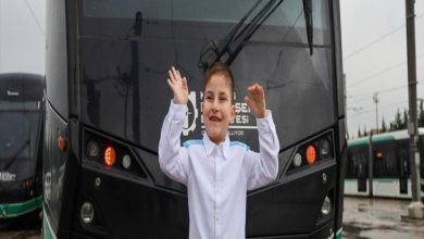 Yağmurlu bir günde, tramvayın önünde duran küçük bir çocuk iki elini havaya kaldırmış, gülümseyerek poz veriyor. Üzerinde beyaz bir gömlek var. Arkada raylı sistem araçları ve şehir altyapısı görünüyor; sahne hem neşeli hem umut dolu bir anı yakalamış gibi.