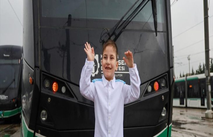 Yağmurlu bir günde, tramvayın önünde duran küçük bir çocuk iki elini havaya kaldırmış, gülümseyerek poz veriyor. Üzerinde beyaz bir gömlek var. Arkada raylı sistem araçları ve şehir altyapısı görünüyor; sahne hem neşeli hem umut dolu bir anı yakalamış gibi.