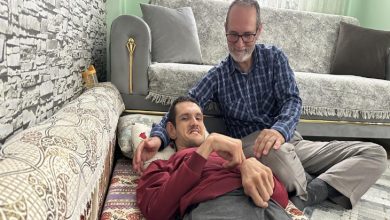 Bir ev ortamında, yerde uzanan genç bir adamın yanında oturan bir kişi ona destek olur gibi omzuna dokunuyor. Arka planda koltuk ve yastıklar yer alıyor. Sahne, aile içi dayanışma ve bakım anını yansıtan sıcak ve samimi bir kare.