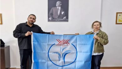 Fotoğrafta bir kadın ve bir erkek, üzerlerinde Yükseköğretim Kurulu (YÖK) logosu bulunan açık mavi bir bayrağı birlikte tutarken görülüyor.