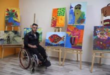 Bedensel engelli bir sanatçının, tekerlekli sandalyesinde oturduğu ve kendi yaptığı rengârenk tablolarla çevrelendiği bir sanat atölyesi karesi. Arka planda şövaleler üzerinde sergilenen balık, çiçek ve portre temalı canlı çalışmalar, atölyenin yaratıcı atmosferini yansıtıyor.