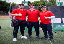 Sahada yan yana duran üç erkek sporcu, kırmızı milli takım formalarıyla poz veriyor. Arkada okçuluk hedefleri görülüyor; sporcular başparmak kaldırarak kameraya gülümsüyor.