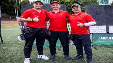 Sahada yan yana duran üç erkek sporcu, kırmızı milli takım formalarıyla poz veriyor. Arkada okçuluk hedefleri görülüyor; sporcular başparmak kaldırarak kameraya gülümsüyor.