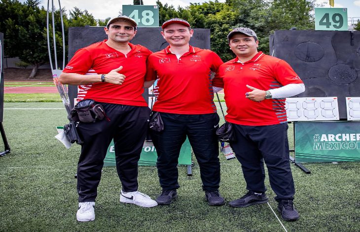Sahada yan yana duran üç erkek sporcu, kırmızı milli takım formalarıyla poz veriyor. Arkada okçuluk hedefleri görülüyor; sporcular başparmak kaldırarak kameraya gülümsüyor.