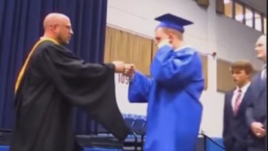 Bir mezuniyet töreni sahnesi: Mavi cübbe ve kep giymiş bir öğrenci, sahnedeki görevliyle yumruk tokuşturuyor. Yan tarafta sırada bekleyen diğer öğrenciler var. Geleneksel tokalaşma yerine daha samimi ve farklı bir kutlama anı yakalanmış.