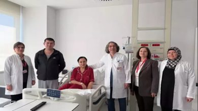 Bir hastane odasında, hasta yatağında oturan bir kadın ve etrafında doktorlar ile sağlık çalışanları birlikte poz veriyor. Ortamda tıbbi cihazlar ve hastane ekipmanları bulunuyor. Sahne, başarılı bir tedavi sonrası çekilmiş, umut ve iyileşme hissi taşıyan bir anı yansıtıyor.