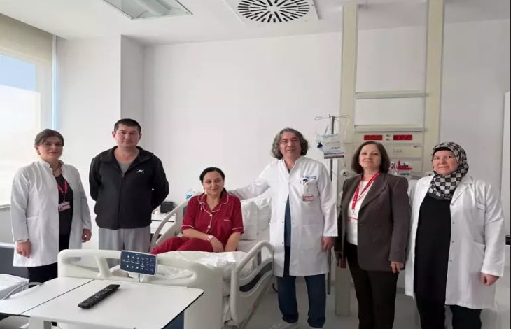 Bir hastane odasında, hasta yatağında oturan bir kadın ve etrafında doktorlar ile sağlık çalışanları birlikte poz veriyor. Ortamda tıbbi cihazlar ve hastane ekipmanları bulunuyor. Sahne, başarılı bir tedavi sonrası çekilmiş, umut ve iyileşme hissi taşıyan bir anı yansıtıyor.