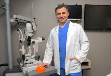 Bir göz muayenehanesinde, beyaz önlüklü bir doktor gelişmiş göz muayene cihazının yanında poz veriyor. Modern tıbbi ekipmanlarla donatılmış ortam, profesyonel ve güven veren bir sağlık hizmeti atmosferi sunuyor.