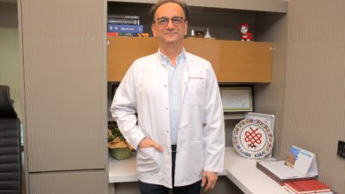 Görselde, bir doktor muayenehane ortamında ayakta duruyor. Beyaz önlük giyen, gözlüklü orta yaş grubunda bir erkek görünüyor. Arkasında kitapların, belgelerin ve dekoratif objelerin bulunduğu raflı bir çalışma alanı yer alıyor. Doktor doğrudan kameraya bakarak sakin bir duruş sergiliyor.