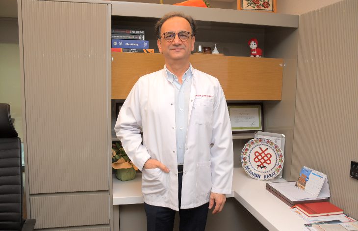 Görselde, bir doktor muayenehane ortamında ayakta duruyor. Beyaz önlük giyen, gözlüklü orta yaş grubunda bir erkek görünüyor. Arkasında kitapların, belgelerin ve dekoratif objelerin bulunduğu raflı bir çalışma alanı yer alıyor. Doktor doğrudan kameraya bakarak sakin bir duruş sergiliyor.