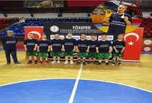 Bir spor salonunda, aynı formayı giymiş bir basketbol takımı saha ortasında yan yana dizilmiş. Arkalarında federasyon logosu ve Türk bayrağı bulunuyor. Görselin köşesinde ise kupayla poz veren bir sporcunun yer aldığı küçük bir ek fotoğraf var. Atmosfer, şampiyonluk gururu ve takım başarısını yansıtıyor.